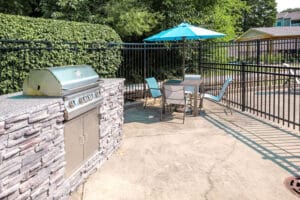 Outdoor patio area with a built-in stainless steel grill, a table with four chairs, and a blue umbrella, surrounded by a black metal fence and greenery.