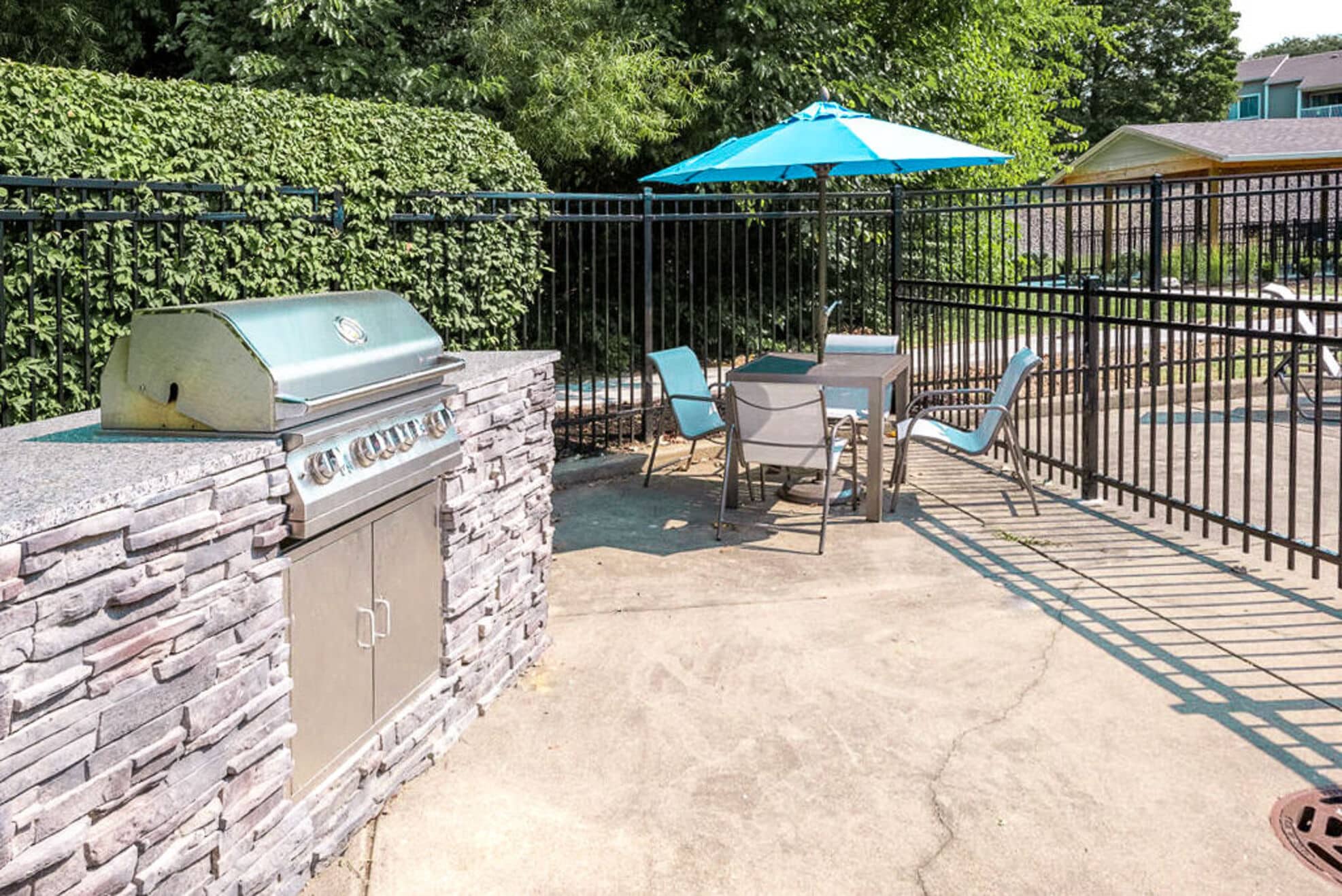 Outdoor patio area with a built-in stainless steel grill, a table with four chairs, and a blue umbrella, surrounded by a black metal fence and greenery.