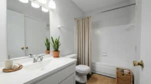 A clean, modern bathroom with a white vanity, mirror, potted plants, toilet, bathtub with beige shower curtain, and wicker baskets on a jute rug.
