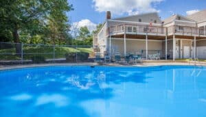 Outdoor swimming pool with clear blue water, surrounded by a fence, patio tables, and chairs, next to a residential building with a wooden deck.