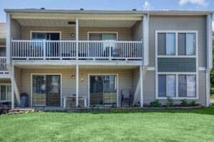 Two-story condo building with beige siding, two balconies, sliding glass doors, and green grass lawn in front.