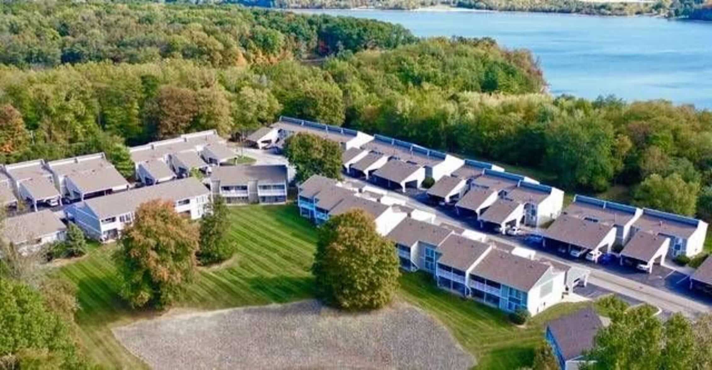 Aerial view of an apartment complex surrounded by trees and greenery, with a lake in the background under a partly cloudy sky.