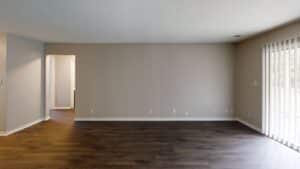 Empty living room with beige walls, dark wood flooring, a doorway leading to another room, and sliding glass doors with vertical blinds on the right side.