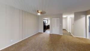 Unfurnished living room with beige walls, carpeted floor, ceiling fan, and view into kitchen and hallway with closets and wood-style flooring.