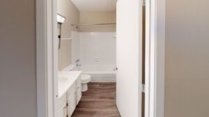 A small bathroom with a white bathtub, toilet, and sink, beige walls, a wooden floor, and an open white door.