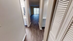 View of a hallway in an apartment with wood-look flooring, leading to a living area with sliding glass doors and natural light. A bathroom and closet are visible on either side.