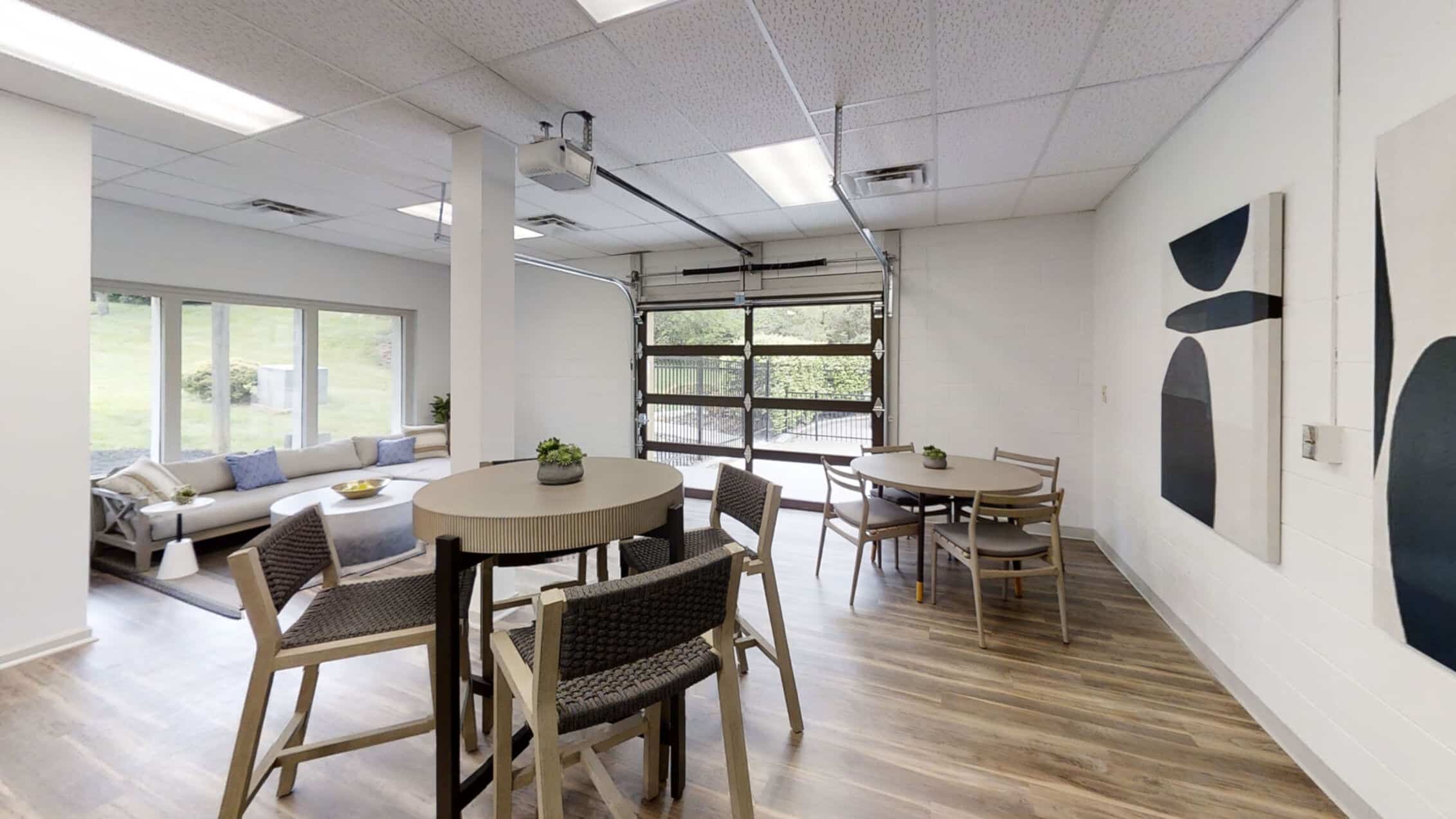 Modern lounge with round tables, chairs, a sofa, large windows, a glass garage-style door, wood flooring, and abstract art on a white wall.