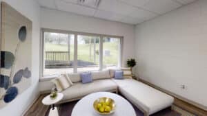 A small lounge area with a white sectional sofa, decorative pillows, a round table with a bowl of lemons, and a window overlooking a grassy yard.