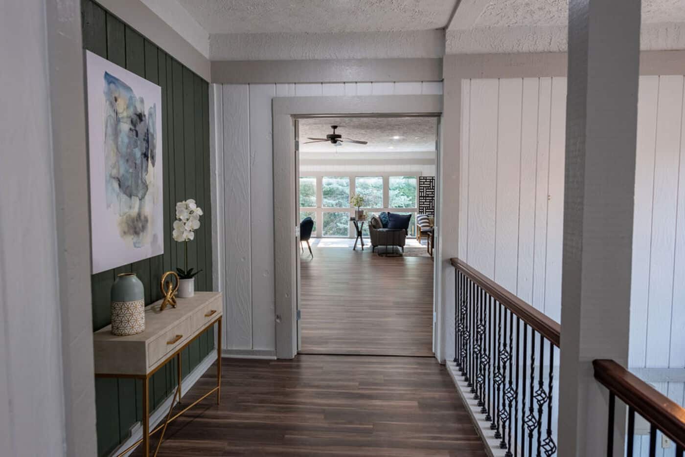 A hallway with wood flooring leads to a bright living room with large windows. A console table with decor and artwork is on the left, and a railing is on the right.