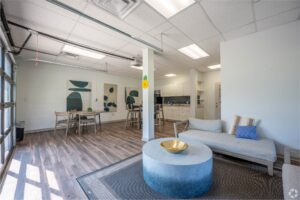 Modern office lounge with a gray sofa, round coffee table, and open dining area; kitchen in the background, art on walls, wood flooring, and bright overhead lighting.