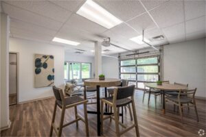 A modern communal room with round tables and chairs, large windows, wood-look flooring, a wall art piece, and a glass garage-style door letting in natural light.