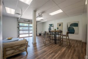 Modern office space with wood flooring, abstract wall art, a high table with chairs, lounge seating, and large glass garage-style door letting in natural light.