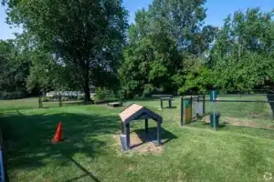 A fenced grassy area with agility equipment, a traffic cone, waste bins, and trees in the background under a clear sky.