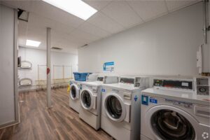 A clean laundromat room with four front-loading washing machines, a laundry basket on top, folding chairs on the wall, benches, and wood-patterned flooring.