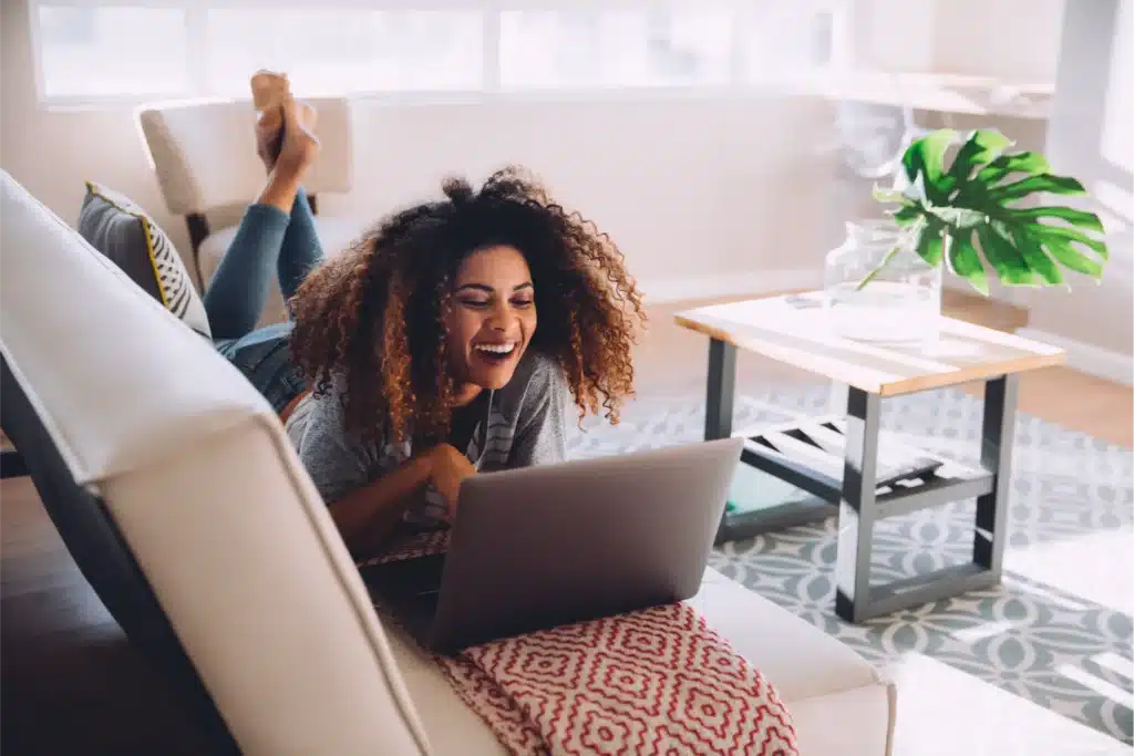 A woman lies on a couch, smiling at a laptop on her lap in a bright living room with a coffee table and a plant.