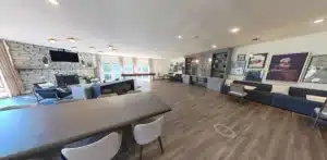Spacious lounge area with wooden floors, modern furniture, stone fireplace, large windows, and artwork on the walls. Tables and chairs are arranged throughout the room.